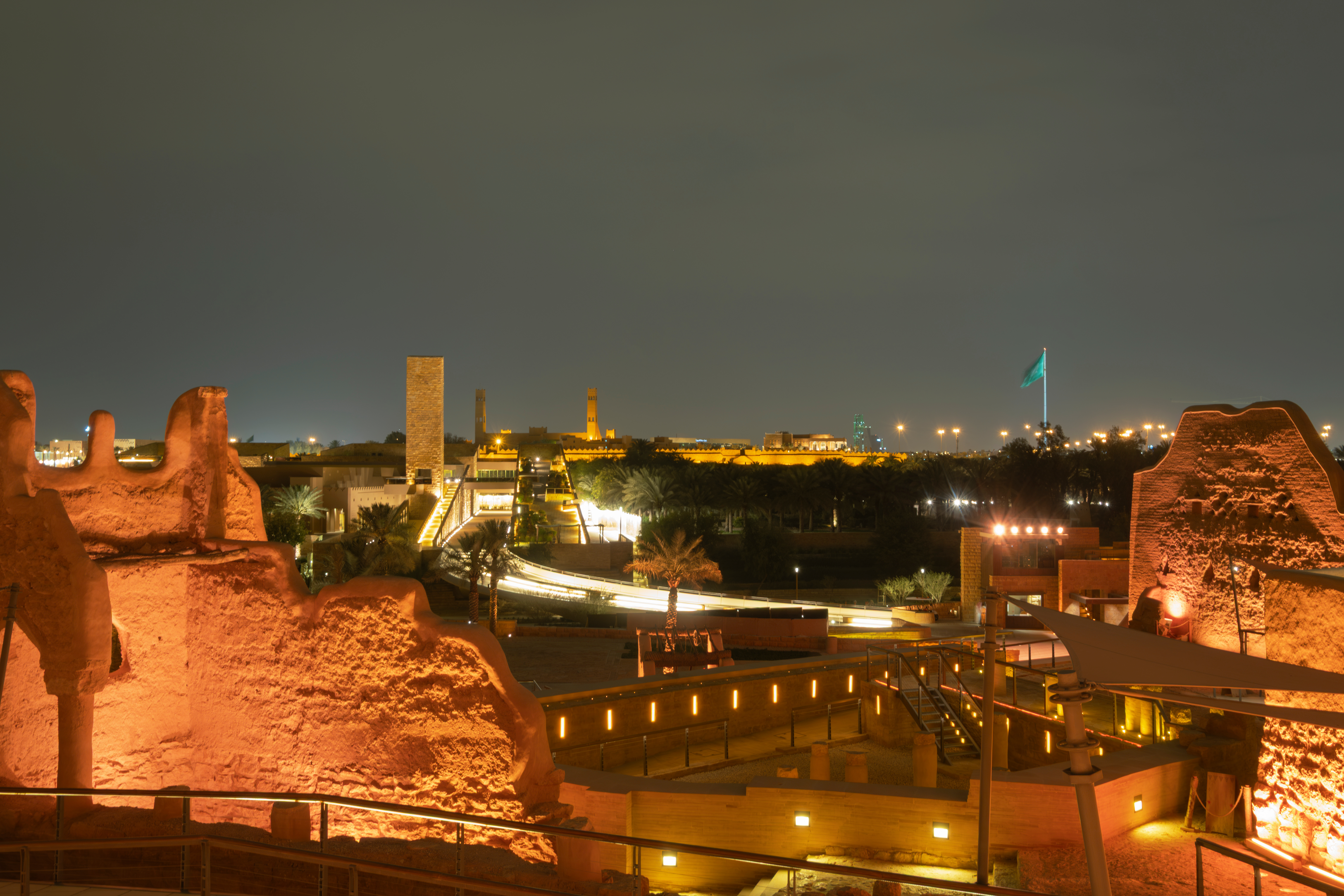 Night view of the illuminated At-Turaif complex in Diriyah, featuring historic mud-brick walls, palm trees, walkways and the Saudi Arabian flag.